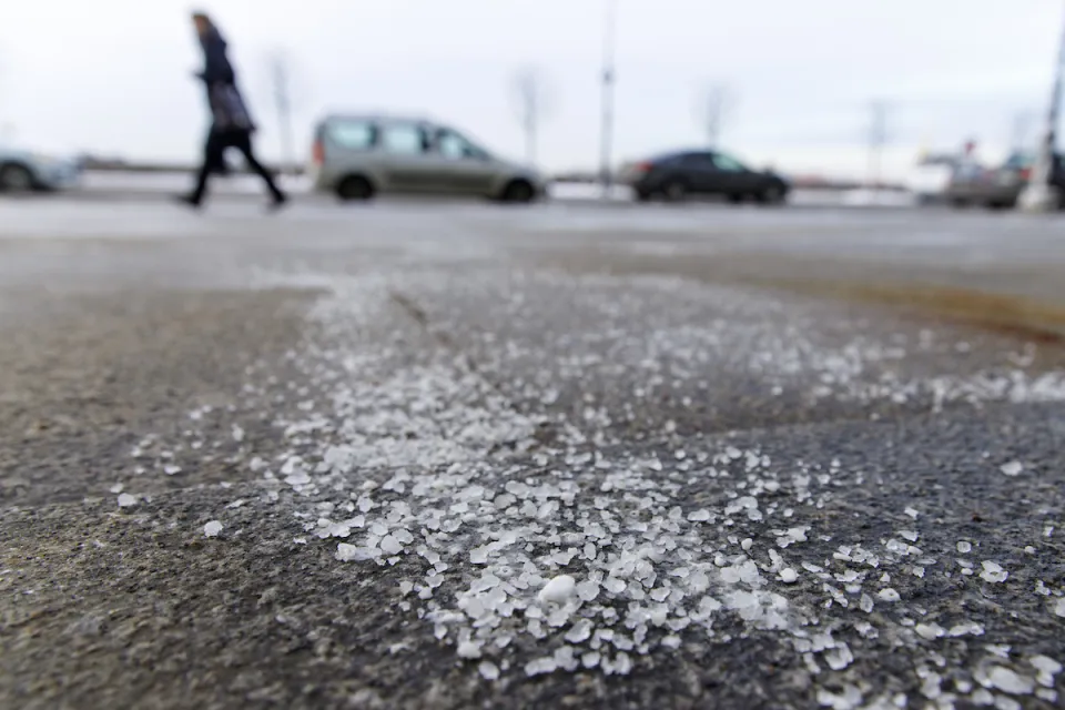 Low close-up of salt sprinkled on asphalt road in winter with person walking and car in background.